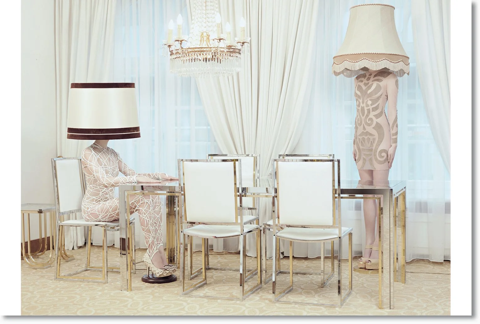 Ein eleganter Raum mit weißen Vorhängen, einem Kronleuchter, einem Couchtisch und einem Esstisch. Es stehen vier Stühle um den Esstisch, zwei davon sind besetzt. Eine Person mit einem Lampenschirm auf dem Kopf sitzt an der Ess-Tafel, eine andere Pers