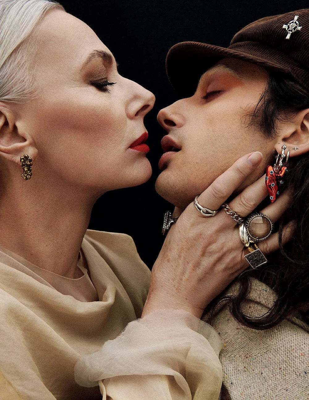 Nahaufnahme eines Pärchens mit engen, verschmolzenen Gesichtern, das sich fast berühren, beide tragen auffällige Schmuckstücke und haben geschlossene Augen.