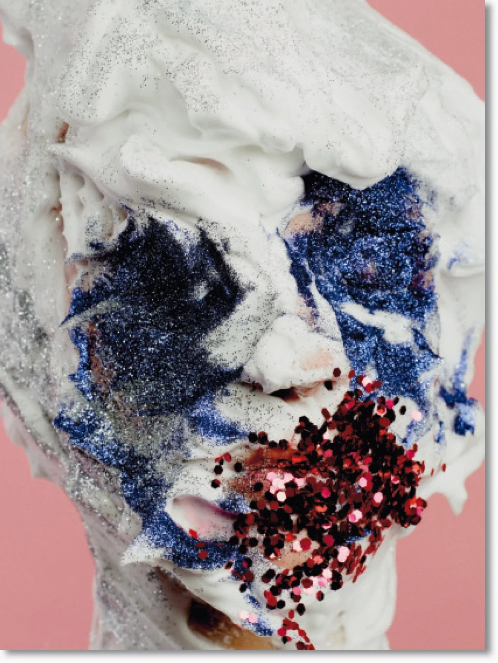 Nahaufnahme einer geschmolzenen Eiswaffel mit weißer Sahne, blauen und roten Glitzer- und Konfettistreuseln auf einem rosa Hintergrund.