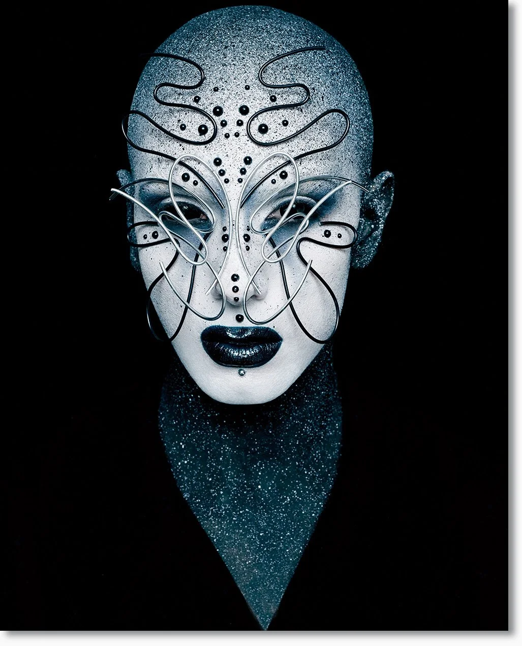 Kunstvolles Maskenporträt einer Person mit schwarzen Lippen, silbernem und schwarzen Make-up, glitzernder Haut und einer komplexen Metallmaske, die das Gesicht überzieht.