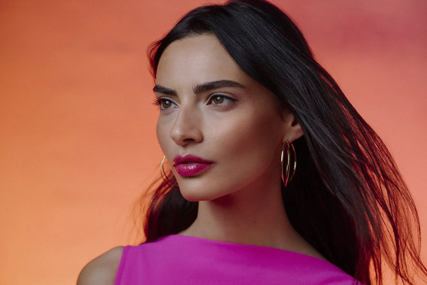 A woman with long dark hair, wearing large gold hoop earrings and a pink top, looking to the side against a warm orange-pink background.