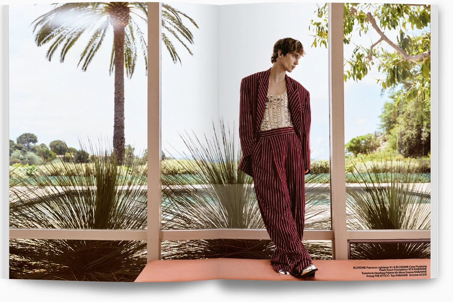 Ein Model steht in einem Raum mit großen Fenstern, durch die man Palmbäume und einen blauen Himmel sieht. Das Model trägt einen roten, gestreiften Anzug mit einer hellen, kurzen Jacke. Die Szene ist hell und erinnert an ein sommerliches Wetter.
