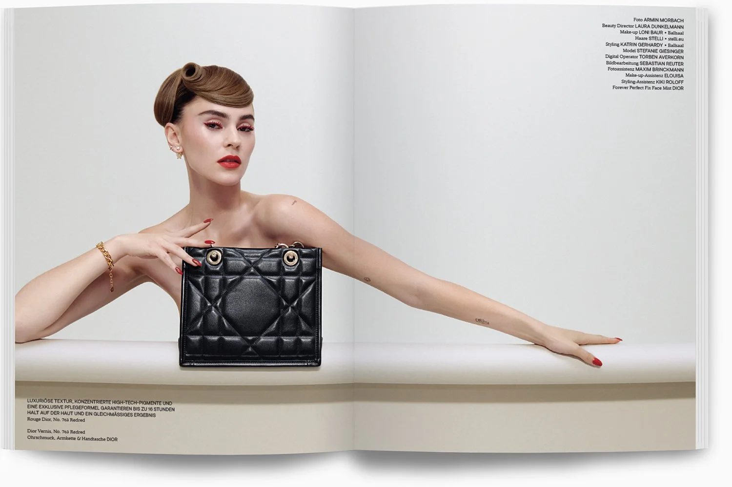 Eine stilvolle Frau mit kurzen, lockigen Haaren sitzt hinter einem weißen Tisch, hält eine schwarze, quilted Tasche von Dior, trägt Goldschmuck und rotes Make-up, vor einem weißen Hintergrund.