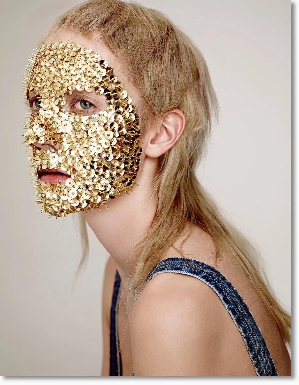 Eine Person mit blonden Haaren trägt eine Maske aus goldfarbenen Nägeln, die das Gesicht verdeckt, nur ein Auge, die Nase und der Mund sind sichtbar.