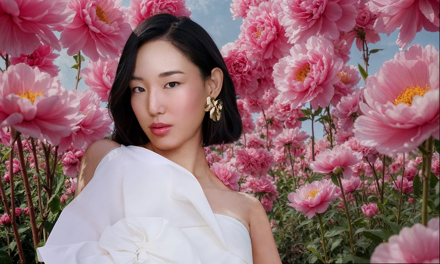 A woman with short black hair wearing a white dress and gold flower earrings, standing among pink peony flowers under a blue sky.