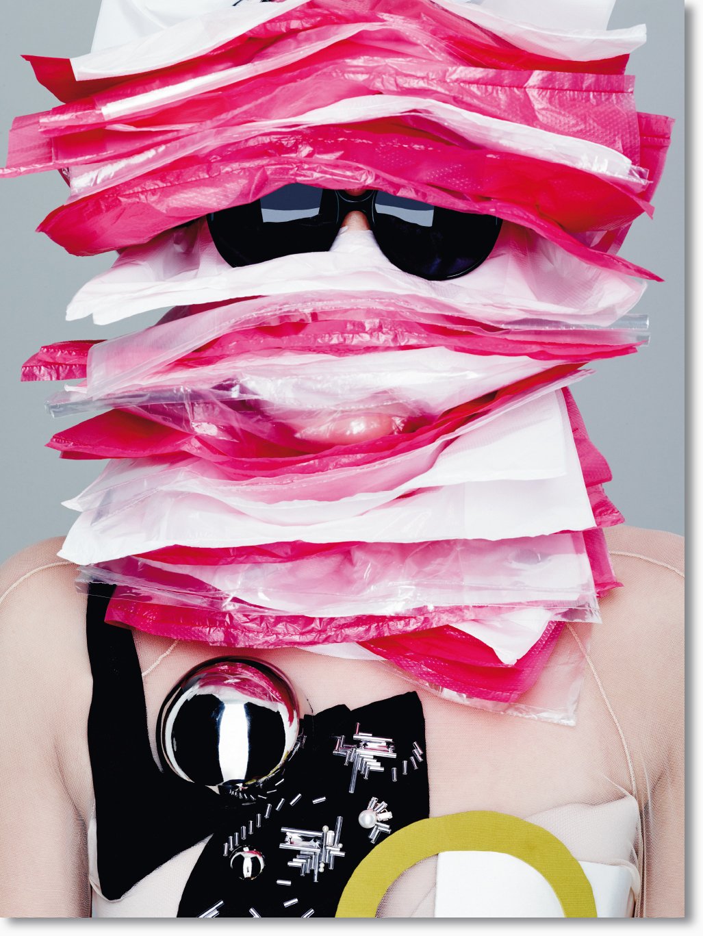 Person trägt eine kreative Maske aus pinken und weißen Plastikbeuteln, Sonnenbrille, schwarz-weiße Kleidung mit metallischen Knöpfen und einem goldenen Ring.