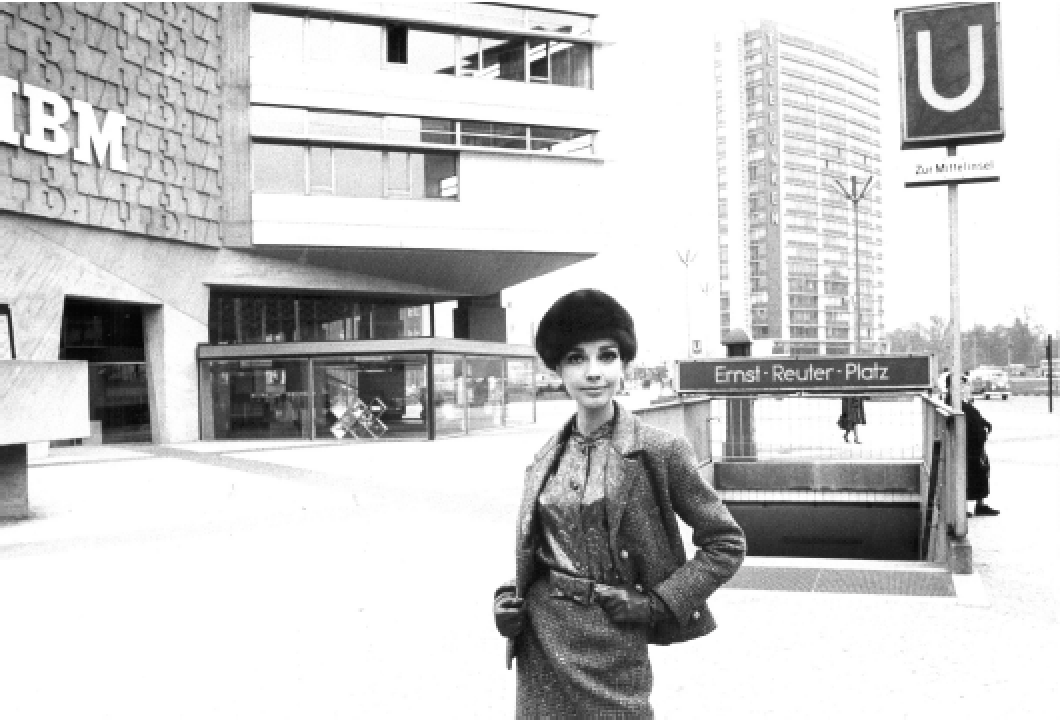 Eine Frau steht vor der U-Bahn-Station Ernst-Reuter-Platz in Berlin, im Hintergrund moderne Gebäude und ein Schild mit der Aufschrift "ZU MITTELS".