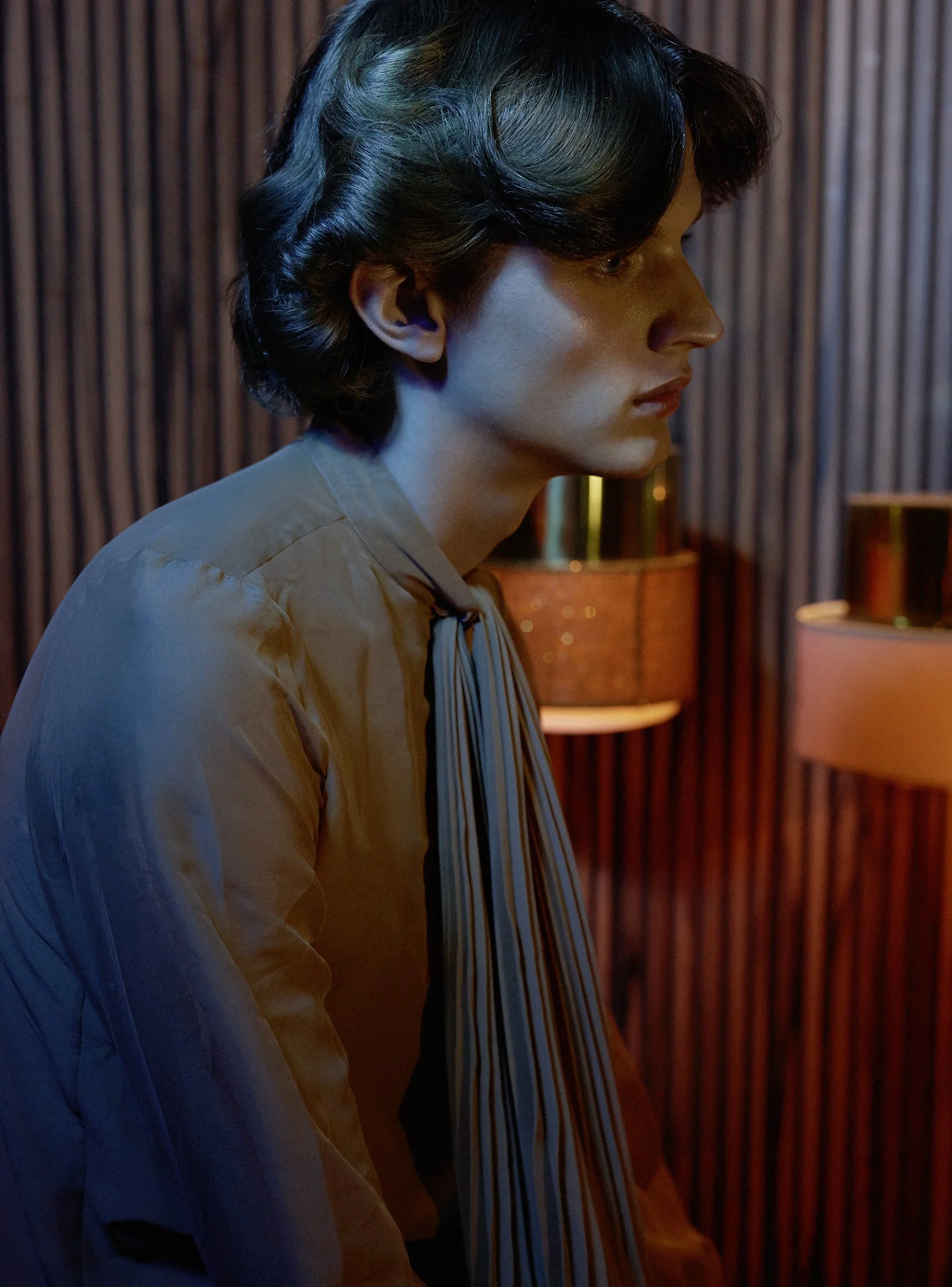 Ein junger Mann sitzt in einem Raum mit dunkler Holzvertäfelung, trägt ein beige Hemd mit Streifen, und ist nach rechts gerichtet, im Profil sichtbar.