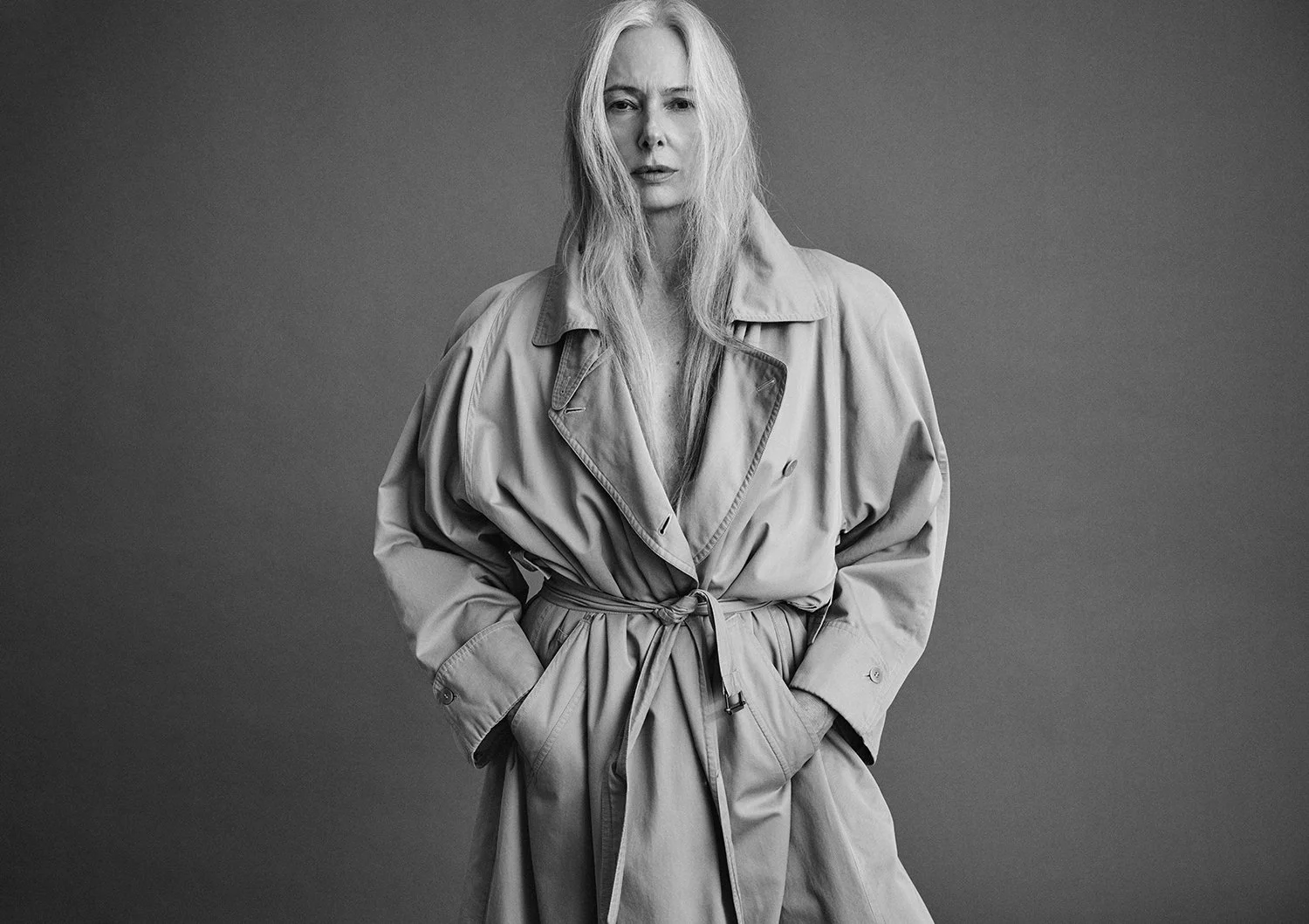 A black-and-white portrait of a woman with long, tousled hair in a trench coat, looking directly at the camera with a serious expression.