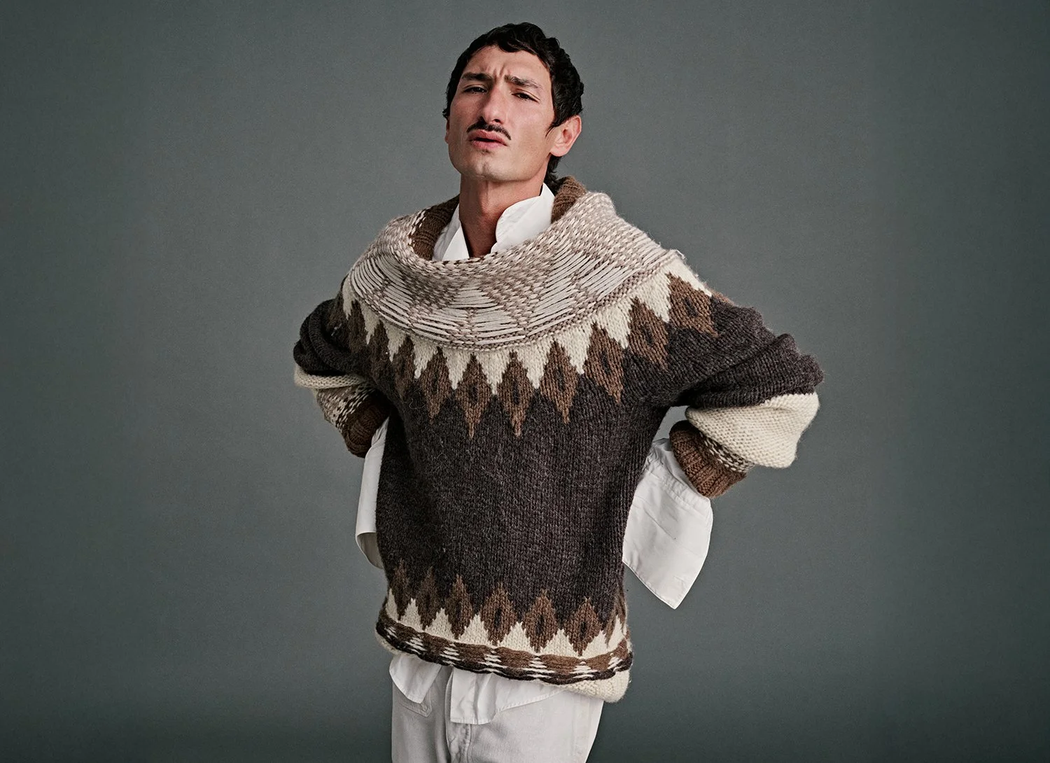 A man with dark hair and a mustache wearing a patterned knit poncho over a white shirt, posing with his hands behind his back against a gray background.