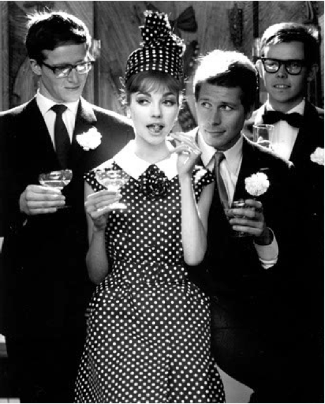 Schwarze und weiße Fotografie von fünf jungen Erwachsenen bei einer Party, alle in festlicher Kleidung, mit Getränken in der Hand, die Frau trägt ein Polka-Dot-Kleid und ein passendes Hut, während die Männer Anzüge mit Fliegen tragen.
