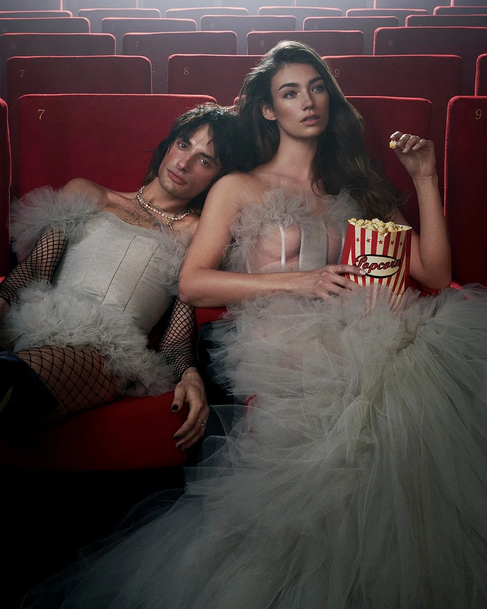 Zwei junge Menschen in glamouröser Kleidung sitzen in einem Kinosaal, einer hält eine Tüte Popcorn, sie sitzen entspannt auf roten Sesseln, umgeben von leeren Sitzreihen.