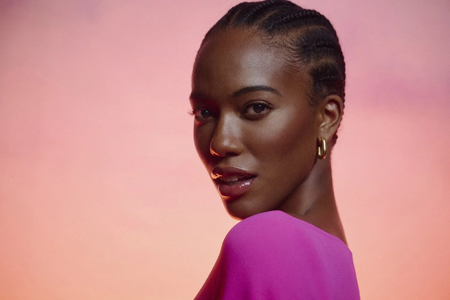 Close-up of a woman with dark skin, braided hair, gold earring, wearing a pink top, against a pink background