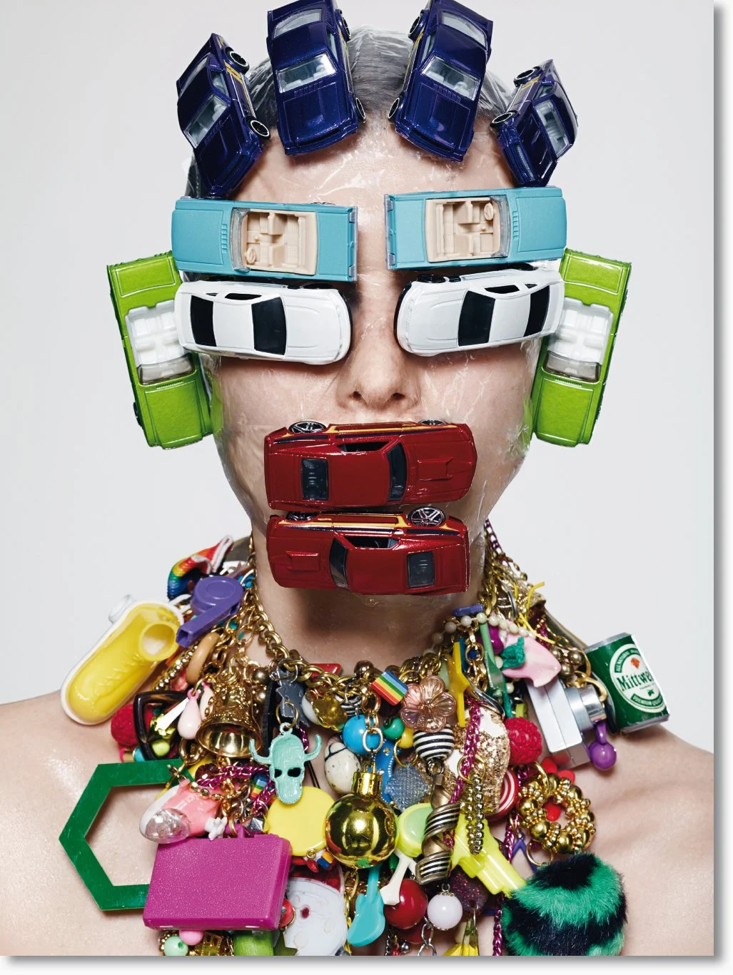 Eine Person mit einer Dusche aus Spielzeugautos, die den Kopf bedecken, trägt große bunte Halsketten und hat Wasser auf der Haut.