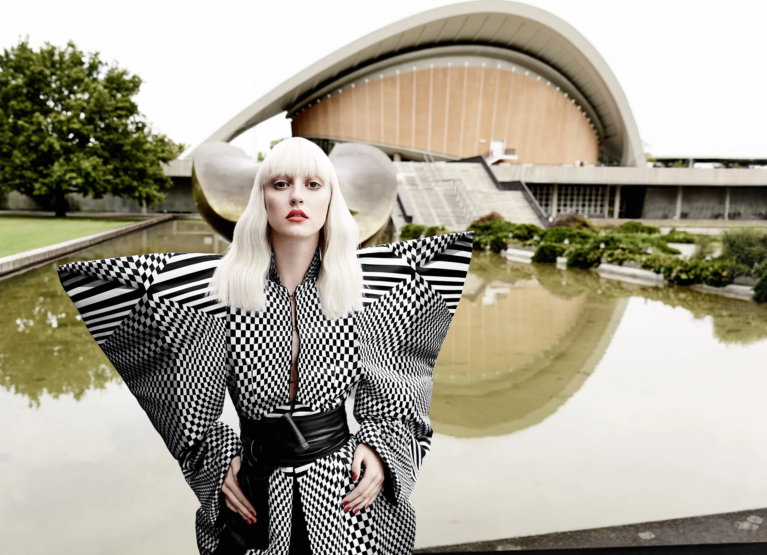 Eine Frau in schwarz-weiß gemustertem Outfit vor moderner Architektur und Wasserfläche.
