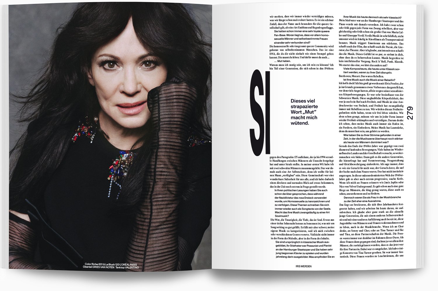 Porträt einer Frau mit schwarzen Haaren, dunklem Make-up und buntem Schmuck, die die Hand vor das Gesicht hält, in einem schwarzen, durchsichtigen Oberteil