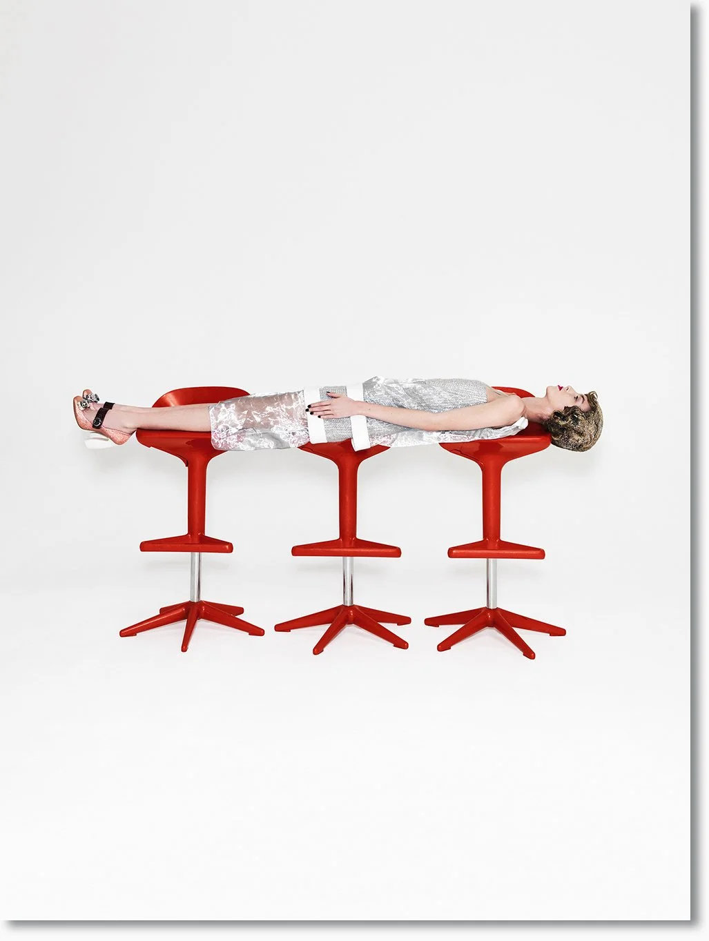 Frau liegend auf drei rote Barhockern, die übereinander gestapelt sind, gegen weißen Hintergrund.