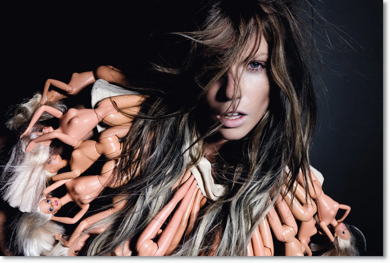 Junge Frau mit langen, gewelltem braunem Haar, umgeben von aufgereihten Barbie-Puppen.