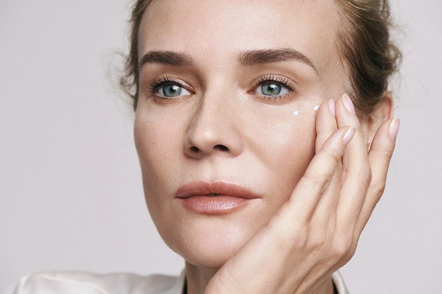 A woman with fair skin and blue eyes applying cream to her face using her fingers.