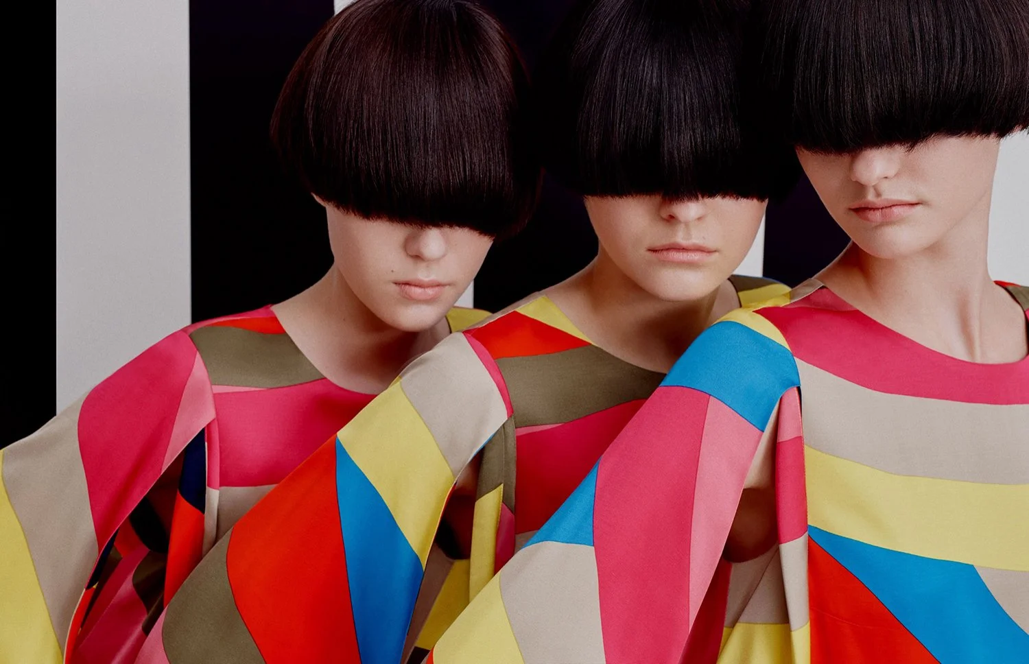 Three women with short dark hair and blunt bangs, wearing colorful geometric-patterned tops, standing close together with their eyes mostly hidden beneath their hair, against a neutral background.