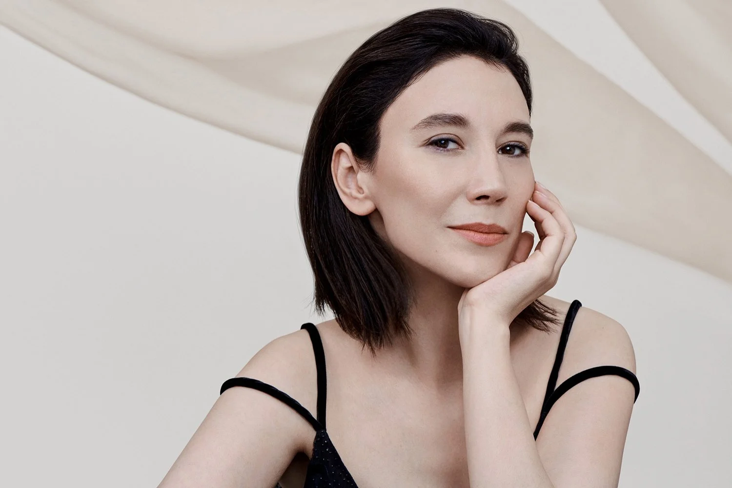 A woman with short dark hair resting her face on her hand, wearing a black spaghetti strap top, against a neutral background.