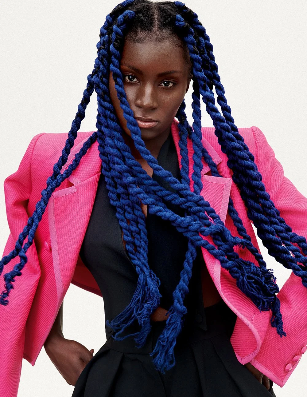 Eine junge Frau mit dunkler Haut, blauen Zöpfen und ernstem Blick trägt eine pinke Jacke und eine schwarze Bluse vor einem weißen Hintergrund.