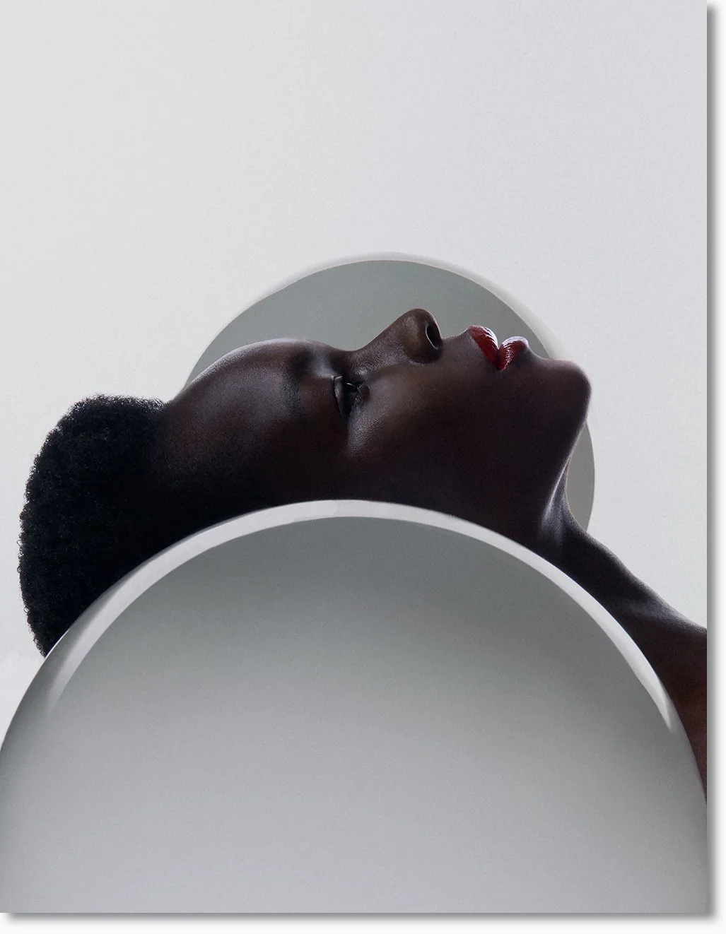 Nahaufnahme eines dunkelhäutigen Frauenkopfs in Profilansicht, der auf einem weißen, runden Objekt liegt, mit roten Lippen und kurzen, lockigen Haaren.