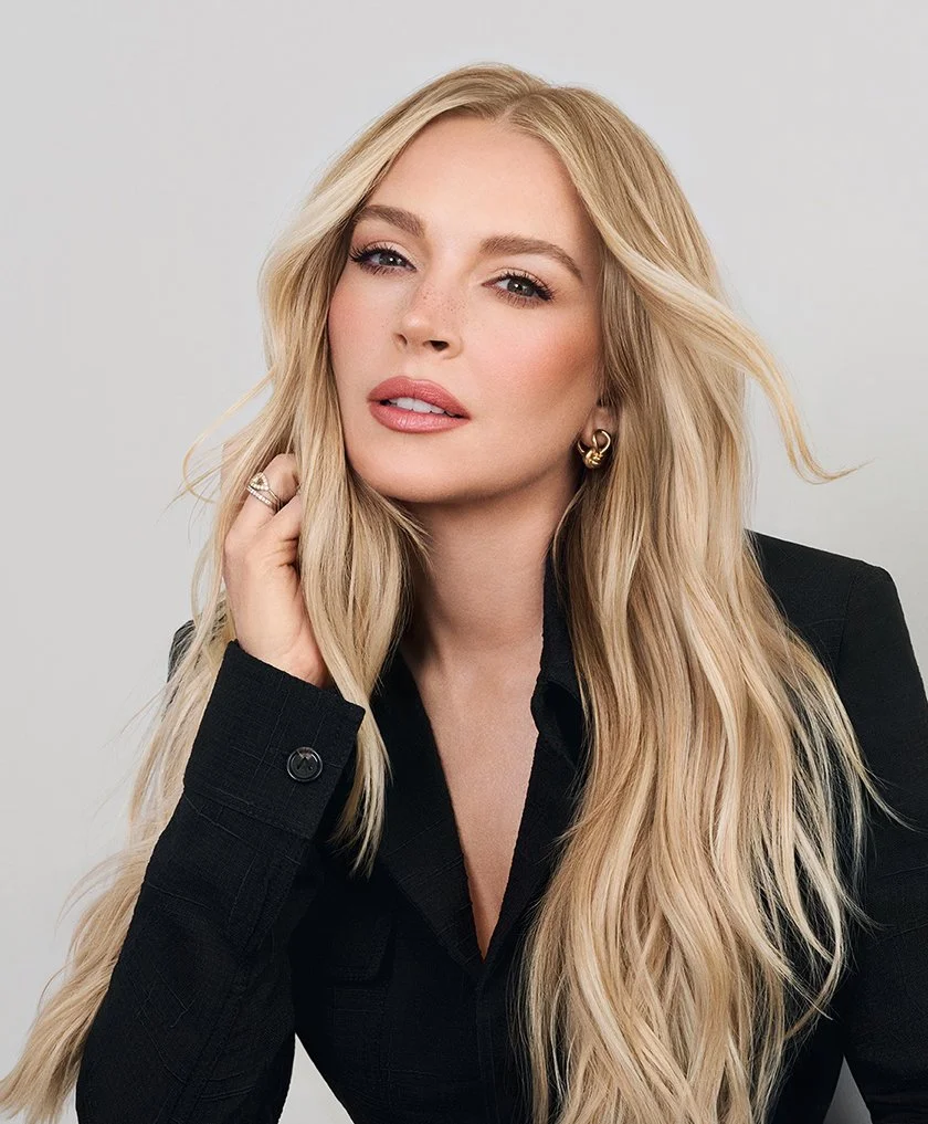 Porträt einer Frau mit langen, blonden Haaren, schwarzer Jacke und Goldohrringen, vor hellem Hintergrund.