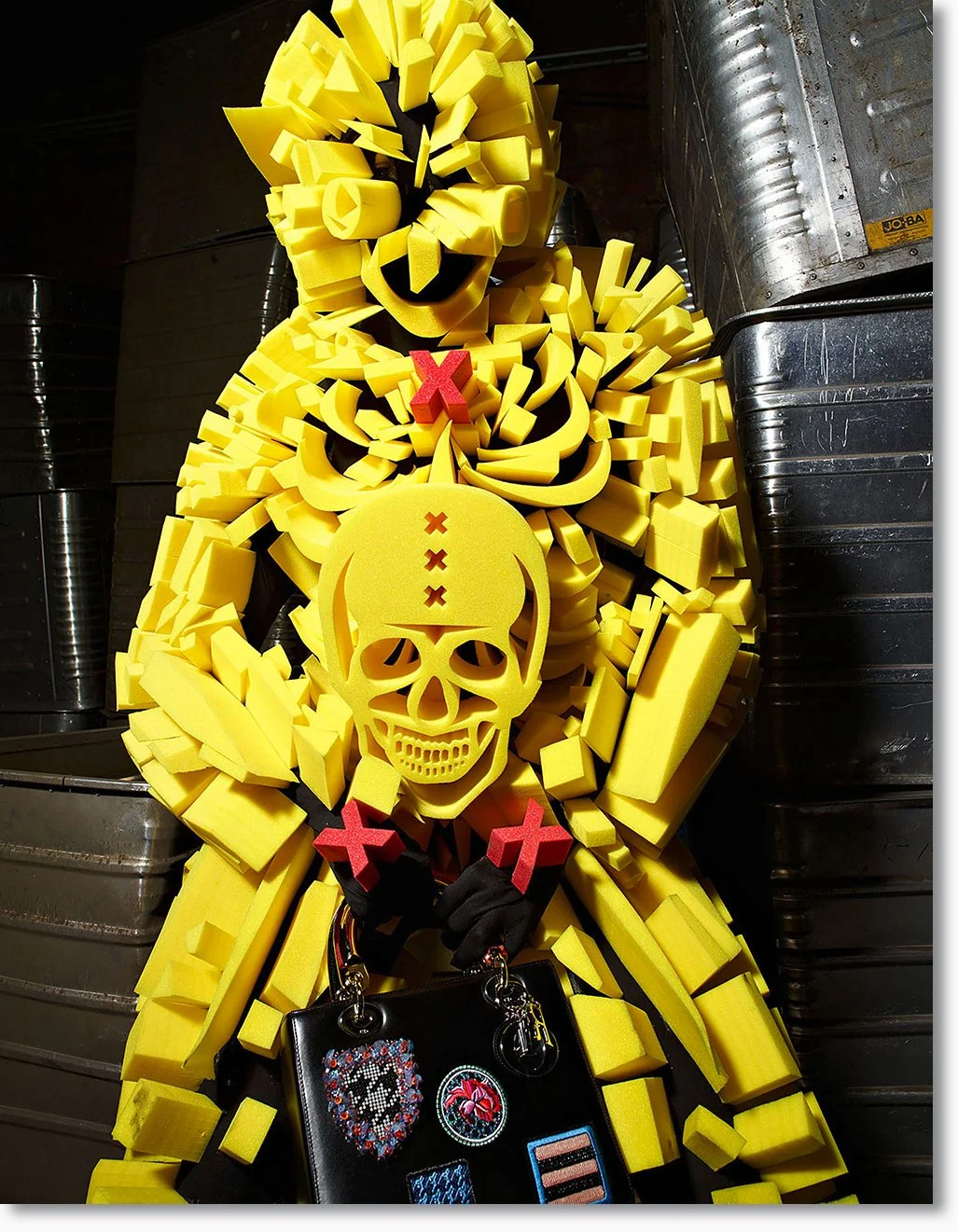 Kunstvolle Skulptur einer Figur aus gelbem Schaumstoff, mit einem Totenschädel und rotem X dekoriert, hält eine Tasche mit Stickereien, umgeben von schwarzen Händen.