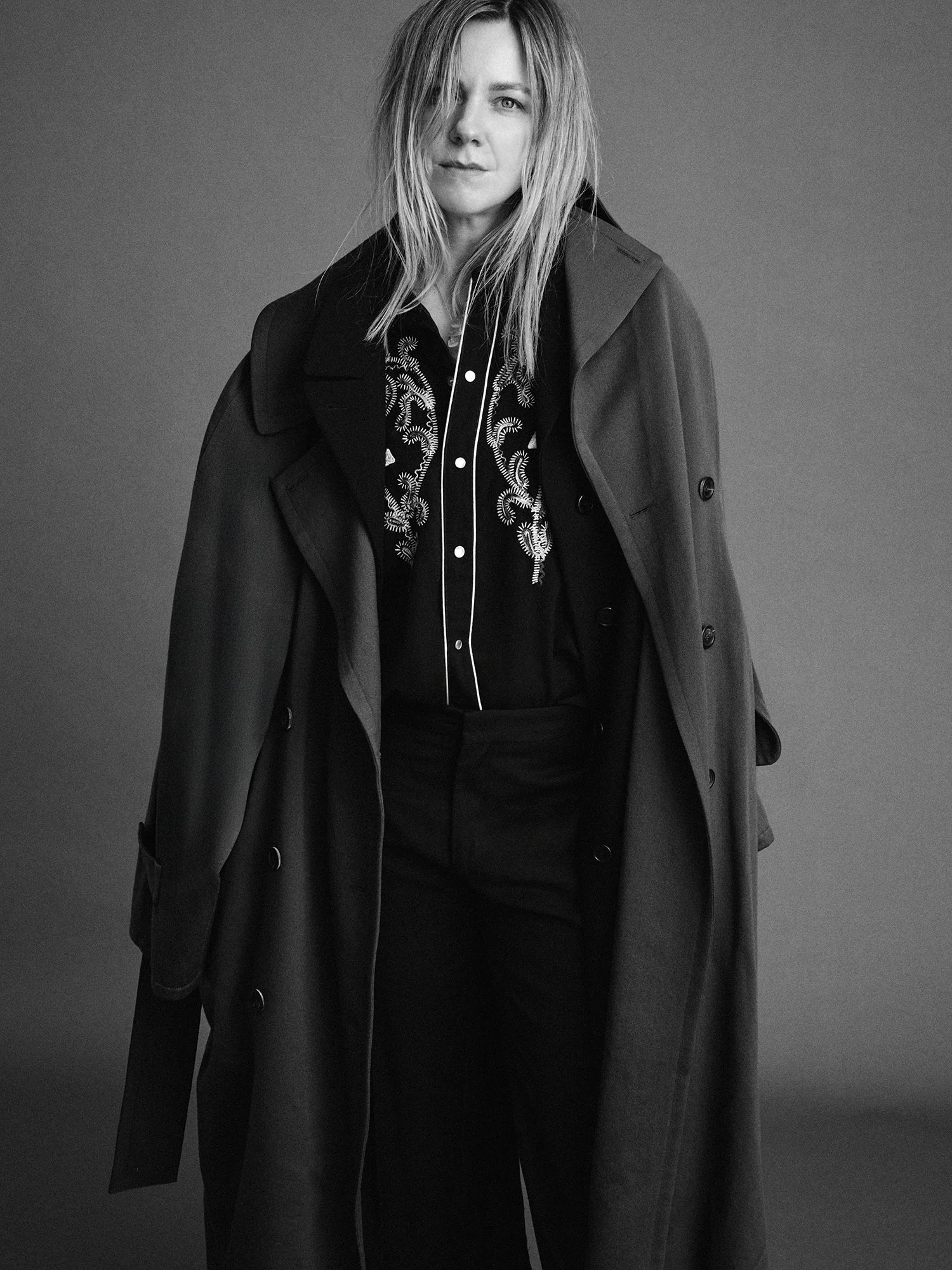 Black and white portrait of a woman wearing an embroidered blouse with a coat draped over her shoulders, standing against a plain background.