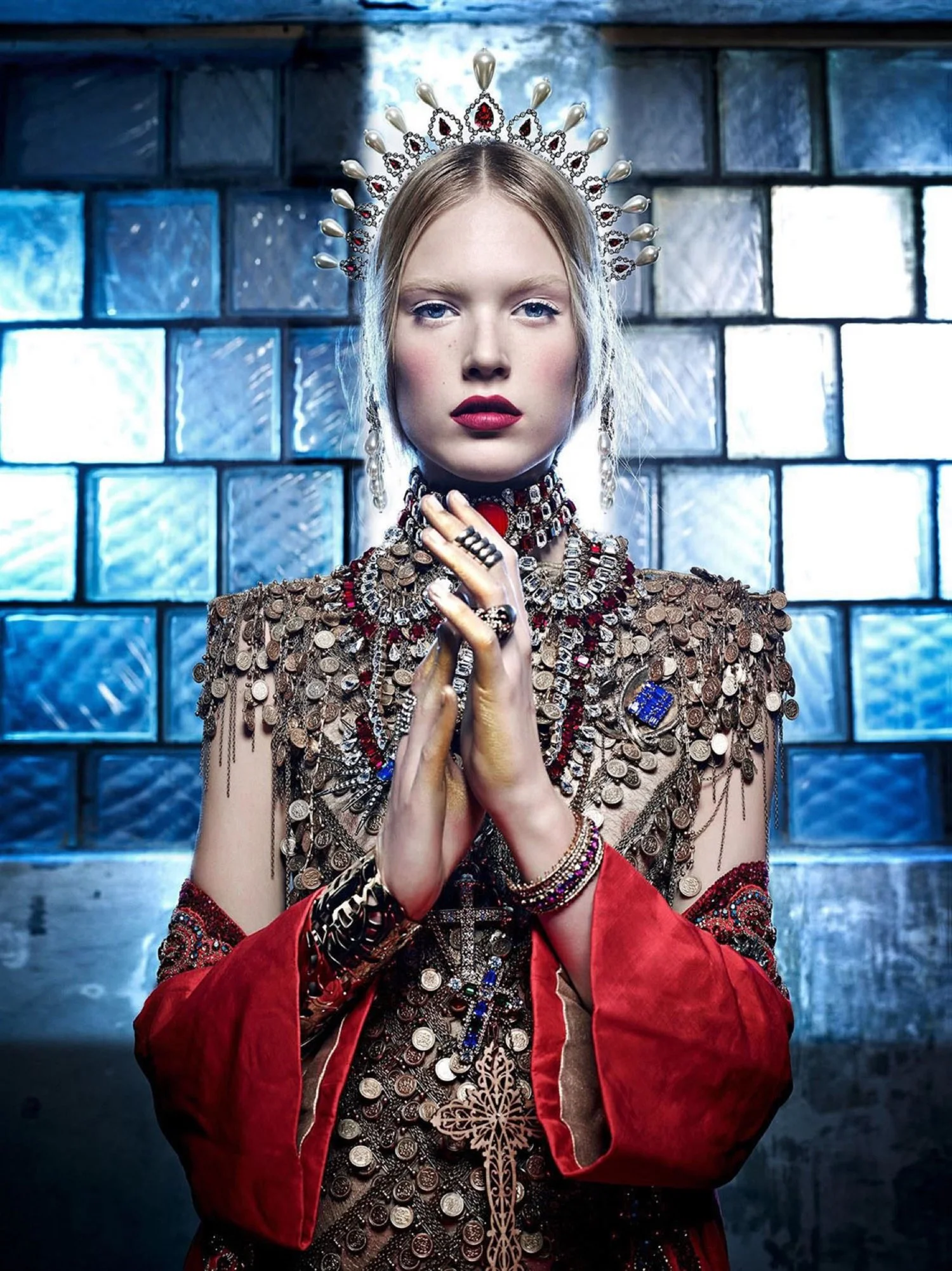 A woman wearing a crown, adorned with jewelry, in front of a blue-tiled wall.
