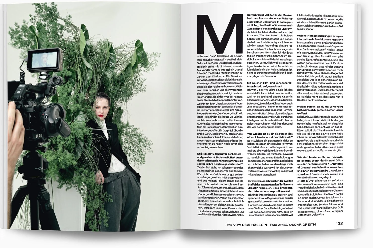 Eine Frau in Schwarz mit weißem Hemd, die neben einer Kamera steht, während im Hintergrund viele grüne Pflanzen stehen. Es scheint sich um ein Fotoshooting zu handeln.