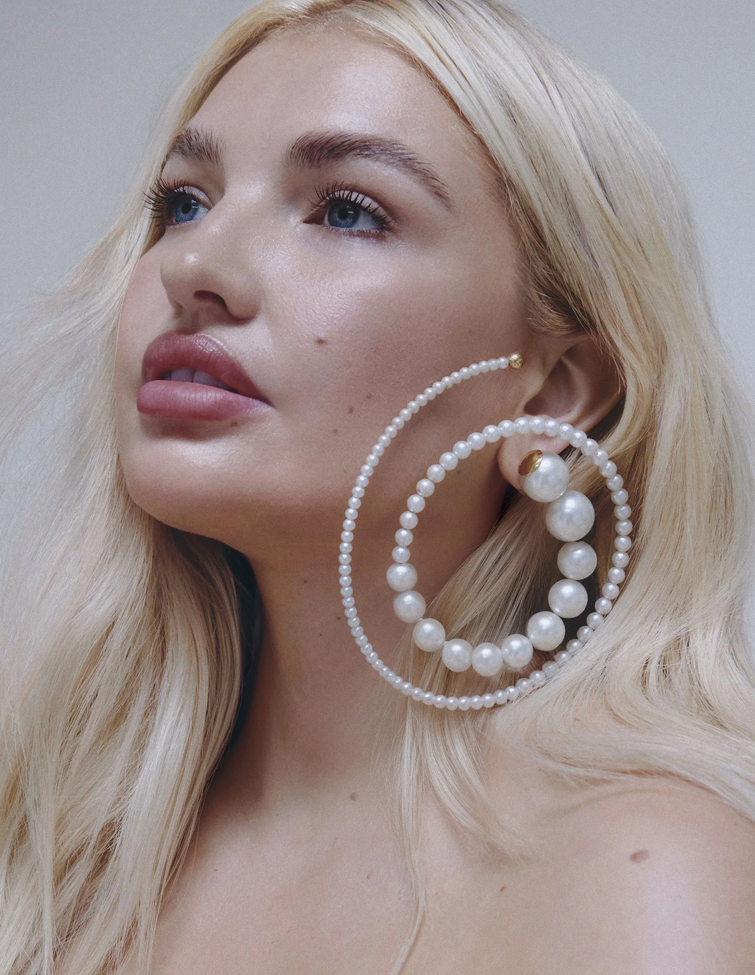 Close-up of a woman with blonde hair and blue eyes wearing large pearl hoop earrings and a pearl necklace, with makeup and natural skin.