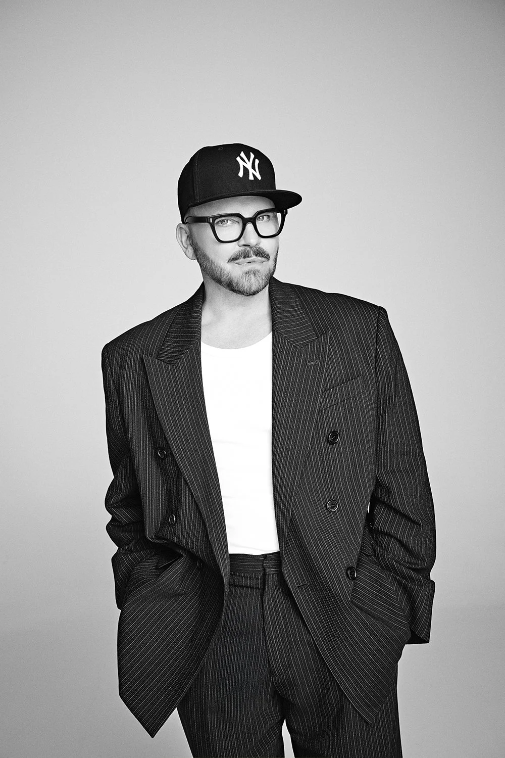 Schwarz-Weiß-Foto eines Mannes mit Brille, sportlicher Baseballcap, Anzug im Nadelstreifen-Muster und weißem T-Shirt, der nach links schaut.