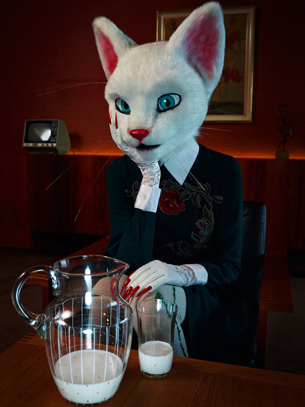 Person mit menschlichem Körper trägt eine große Katze-Maske, sitzt an einem Tisch mit Milch, im Hintergrund eine Wand mit Gemälde und einem Fernseher.