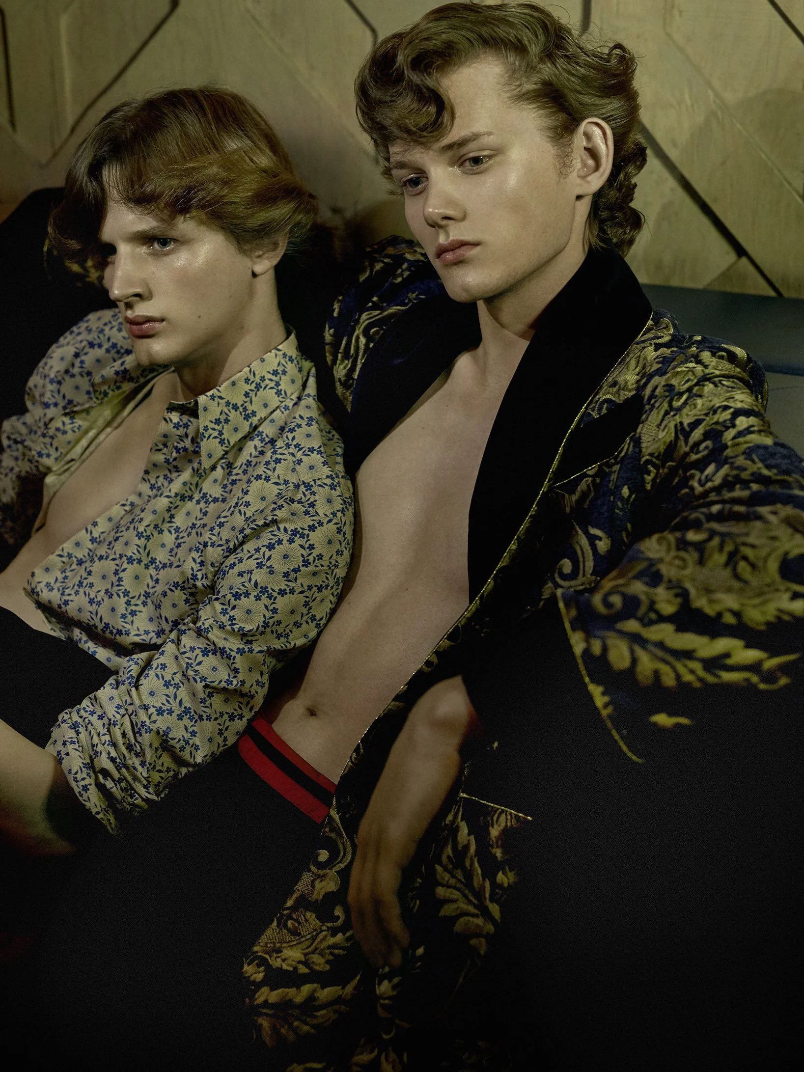 Zwei junge Männer sitzen auf einer Couch, einer trägt ein offenes, elegantes Jackett, der andere ein Hemd mit Blumenmuster. Beide schauen nach unten und wirken nachdenklich.