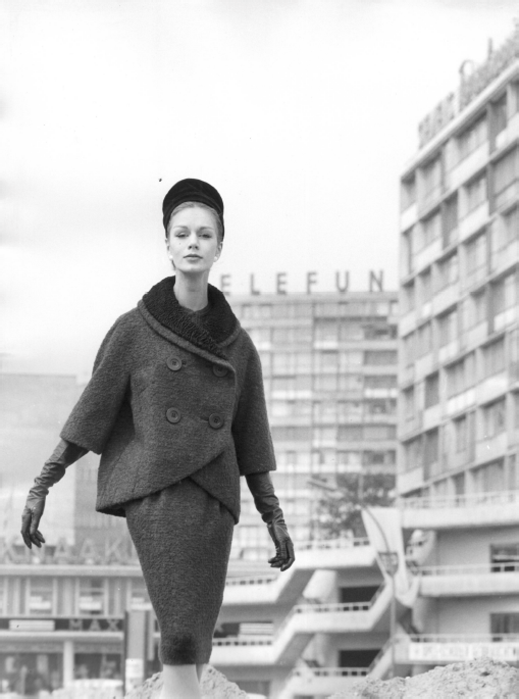 Schwarz-Weiß-Fotografie einer eleganten Frau in Vintage-Kleidung, die in einer Stadt steht, im Hintergrund ein Gebäude mit dem namensschild "ELEFUNK".