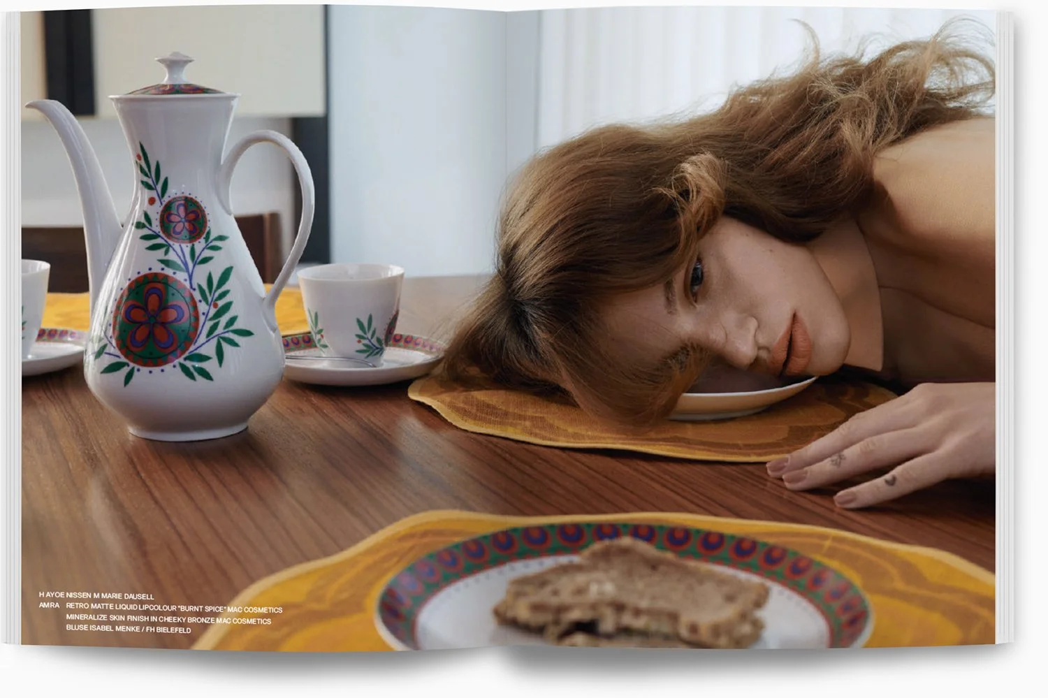 Frau liegt mit Kopf auf einem Teller auf einem Holztisch, umgeben von buntem Geschirr und einer Teekanne, während sie einen Kaffee hat und eine Scheibe Kuchen vor sich liegt.