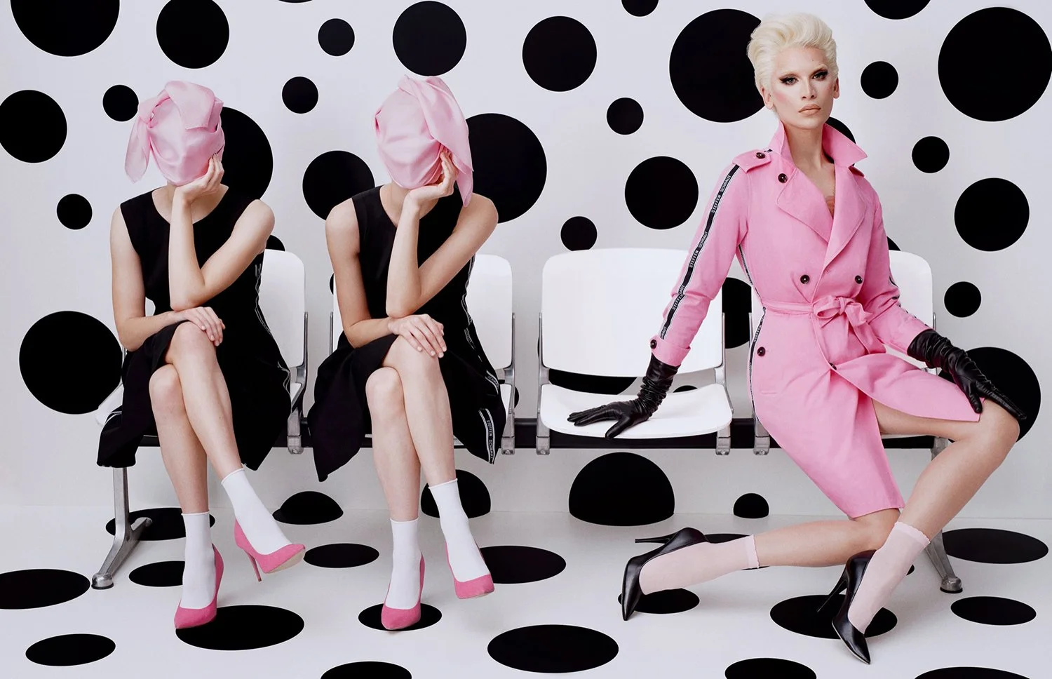 Four women sitting on white chairs against a white wall with black polka dots; two women have pink headscarves and black dresses, one woman is in a pink trench coat with black gloves, black high heels, and light stockings.