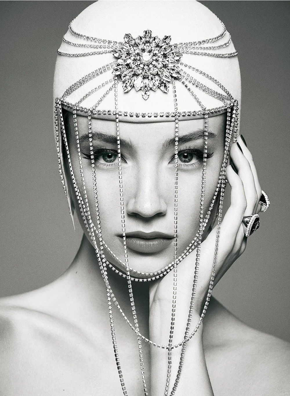 Schwarz-weiß-Fotografie eines Models mit kunstvollem Kopfstück aus funkelnden Ketten und einem großen Kristall-Blumen-Emblem, das wie ein Diadem auf dem Kopf sitzt, mit der Hand an der Seite des Gesichts.