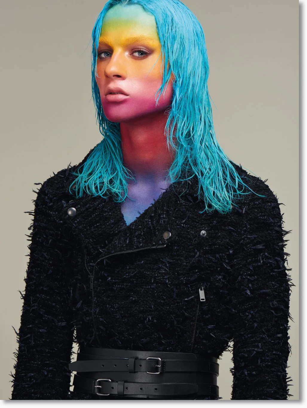 Junge Frau mit nassen, blauer, gewellter Haarfrisur und multicoloren Gesichtsbemalung in Pink, Gelb, Grün und Blau, trägt schwarzen, spitzen Textur-Overall mit Gürtel.