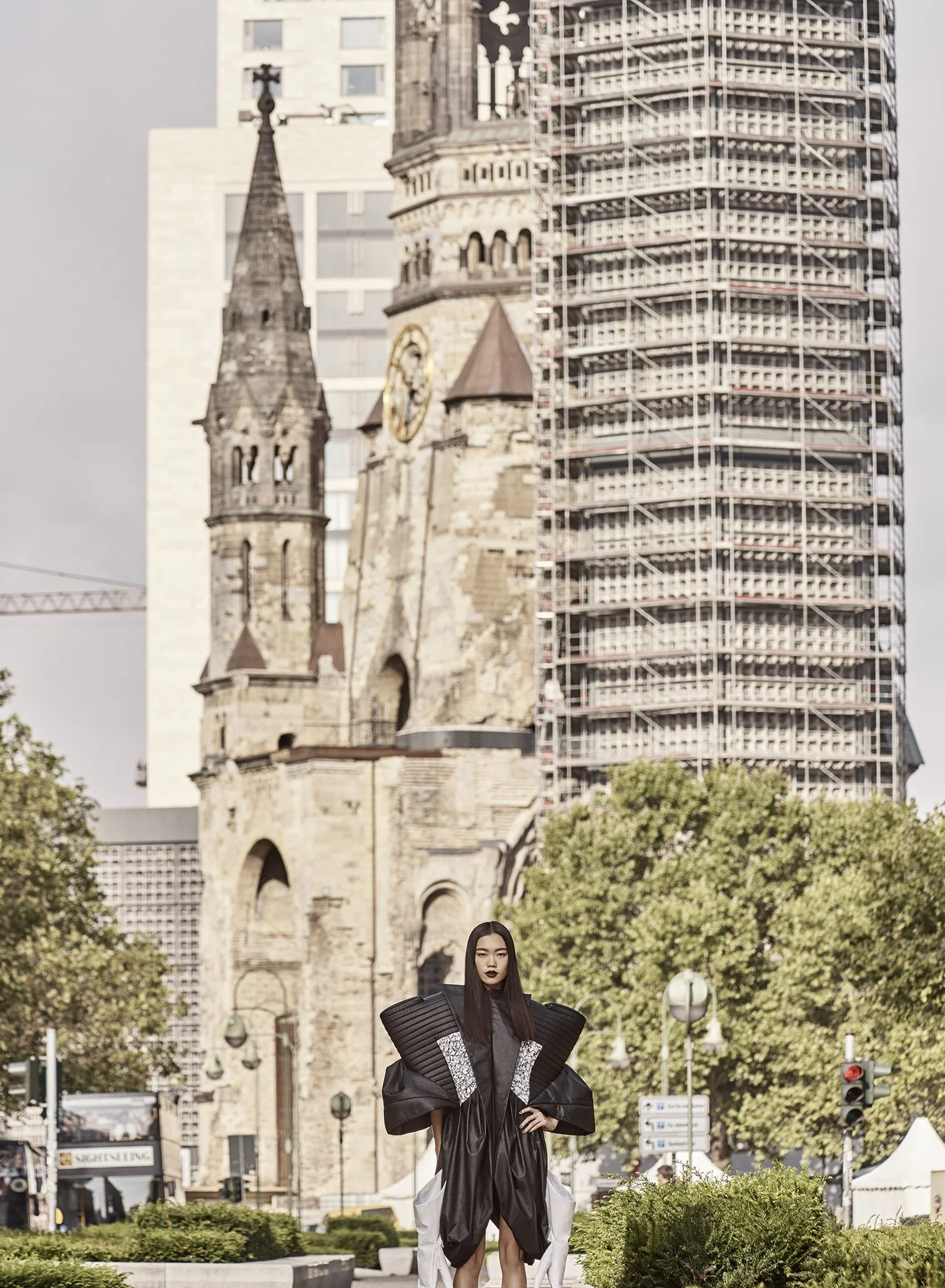 Eine Frau in auffälliger schwarzer Mode mit großen Schulterstücken steht vor der Kirche St. Sebastian in einem städtischen Bereich, umgeben von Bäumen, Baustellen und moderner Architektur.