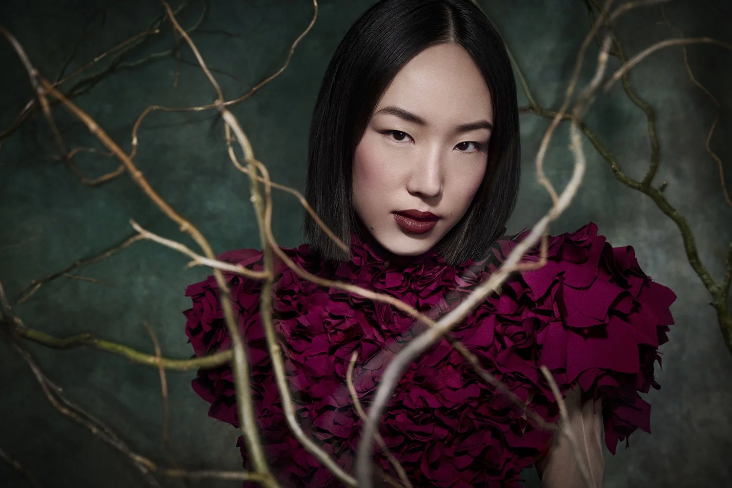A woman with short black hair and dark lipstick looking at camera, wearing a deep red ruffled top. She is surrounded by twisting, dried branches with a dark green background.