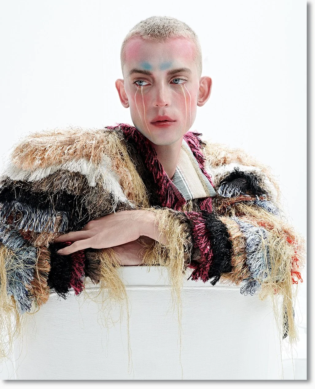 Porträt eines jungen Mannes mit regenerierter, kurzen, blonder Frisur, makeup-artikoulisiertem Gesicht und außergewöhnlicher, bunten, pelzartigen Jacke auf weißem Hintergrund.