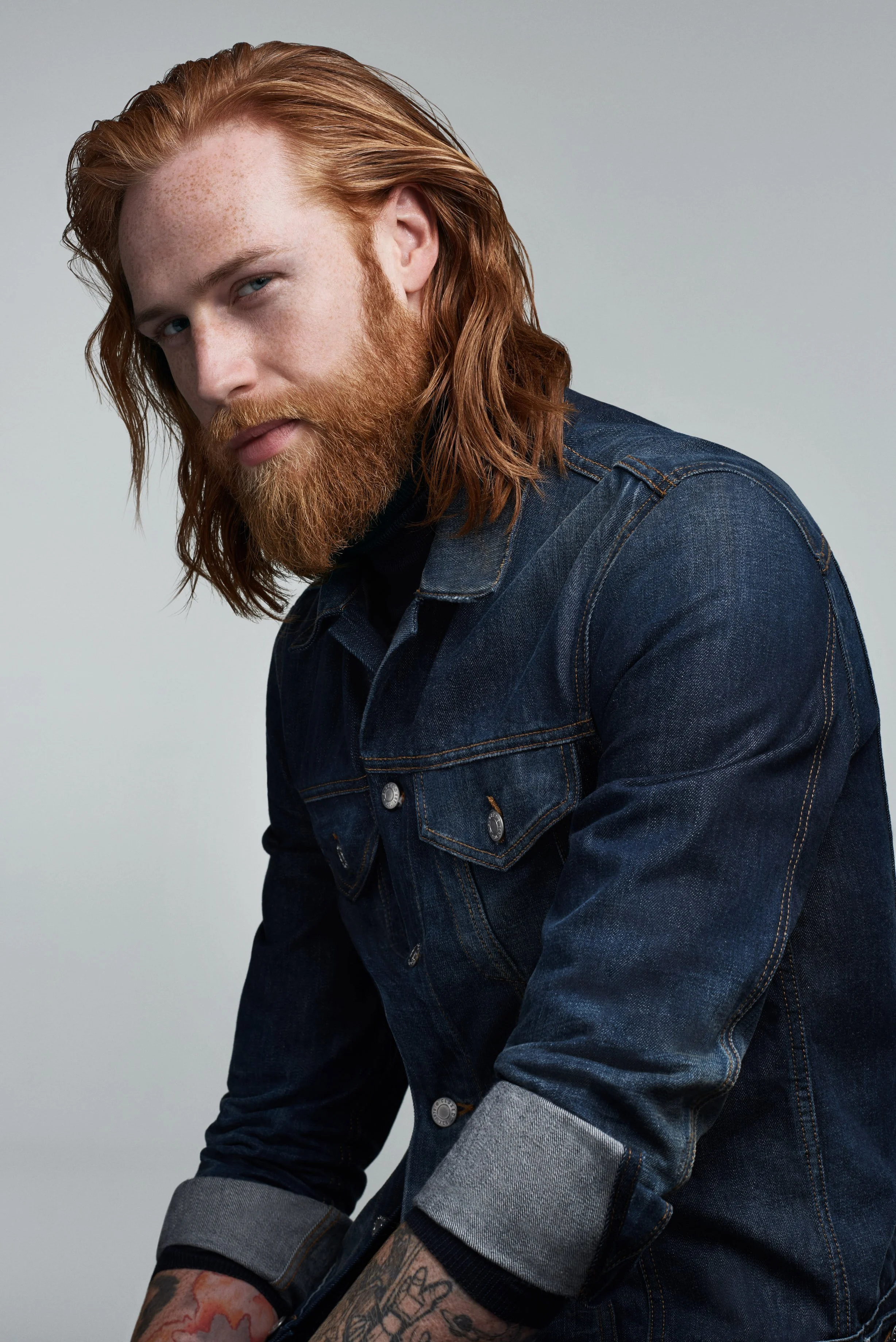 Portrait eines Mannes mit roten Haaren und Bart, in Jeansjacke, in neutralem Hintergrund.