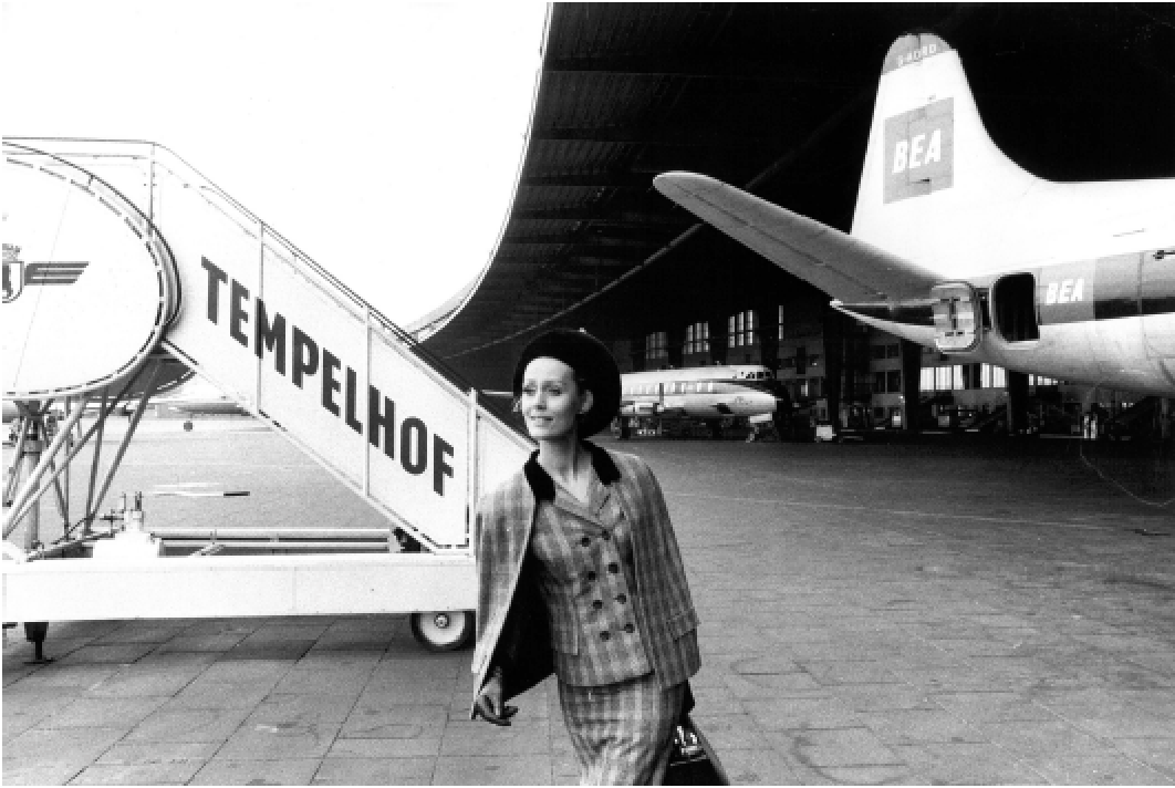 Eine Frau in einem karierten Anzug und Hut steht vor einem Flugzeug und einer Treppe mit dem Schriftzug 'TEMPELHOF'.