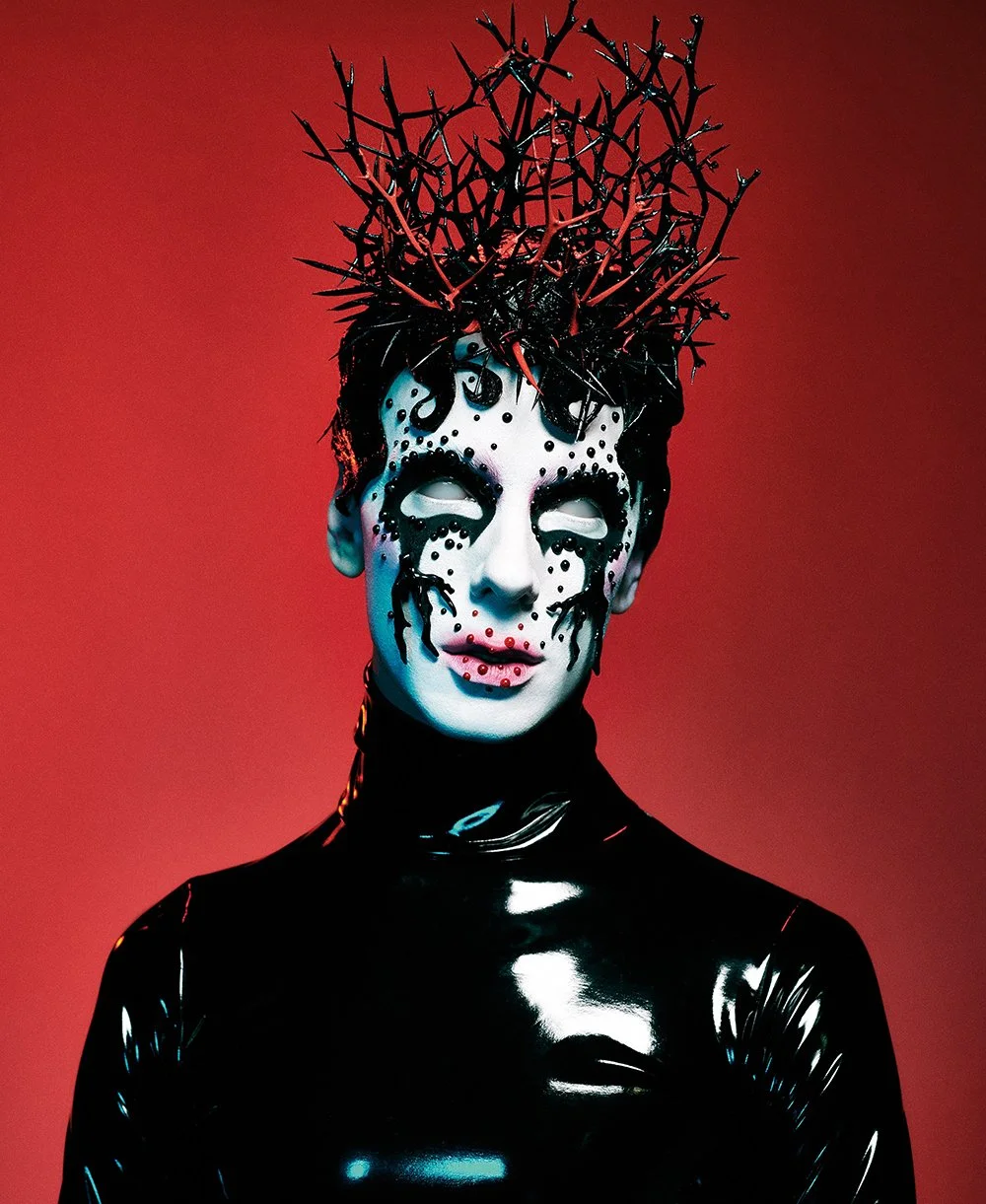 Porzellan-ähnliches Gesicht mit schwarzen und roten dekorativen Elementen, getragen von einer Person in einem schwarzen, glanzenden, engen Outfit, gegen roten Hintergrund, mit einer Dornenkrone aus roten Zweigen.