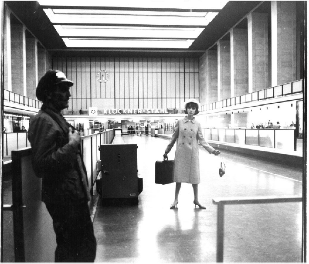 Zwei Menschen in einem Flughafen, einer männlich und eine weibliche Person. Die männliche Person steht links im Vordergrund mit einer Hand an der Brust, die weibliche Person steht in der Mitte mit Koffer in der Hand. Die Architektur des Flughafens is