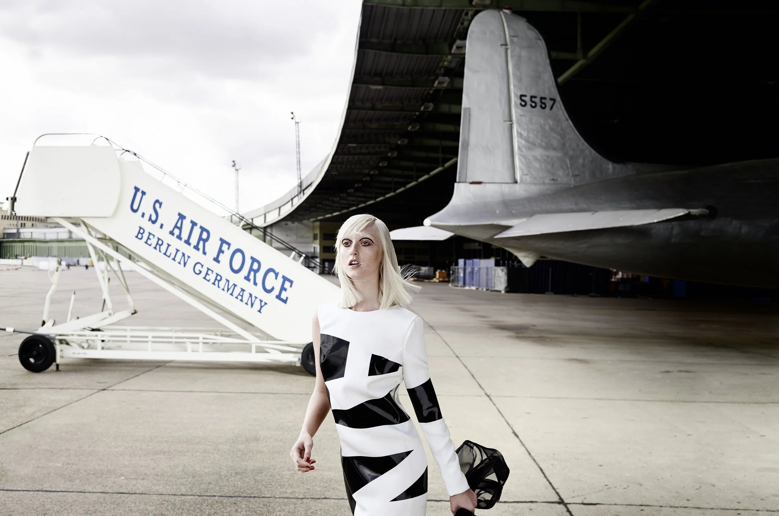 Frau mit blondem Haar in einem schwarzen-weißen Kleid im Flughafen, im Hintergrund ein Flugzeug und eine Treppe mit der Aufschrift U.S. AIR FORCE, BERLIN GERMANY.