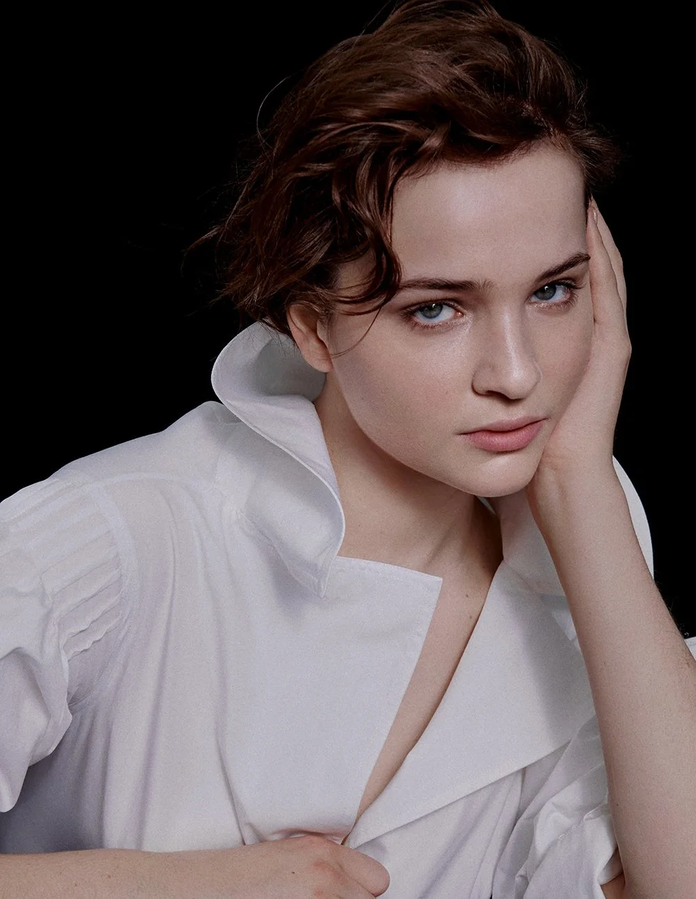 Porträt eines jungen Mädchens mit braunen, lockigen Haaren und blauen Augen, trägt einen weißen Hemd-Kragen und schaut direkt in die Kamera, steht vor schwarzem Hintergrund.